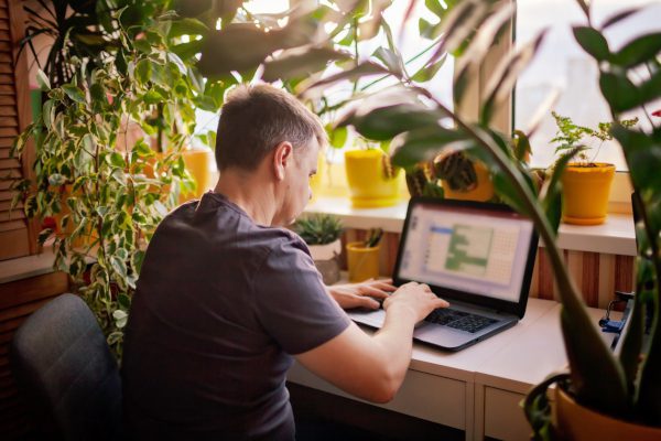 A man working with laptop remotely from home. A distant work place with many home plants. Green nature inspired home office. Indoor lifestyle