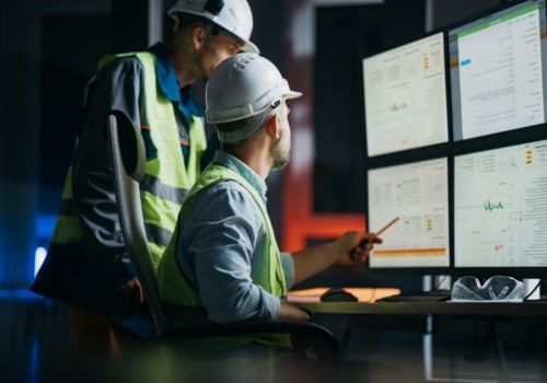 Two men in safety gear overlooking data on monitors