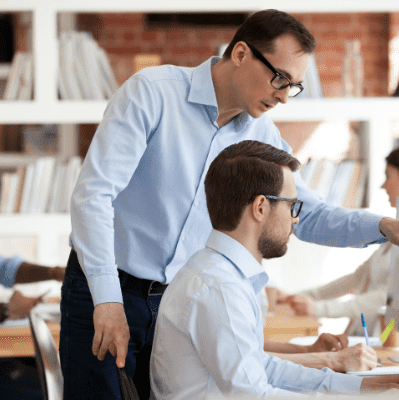 office setting with one man standing over another man at a desk pointing to a monitor